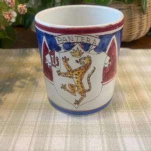 May include: A white ceramic mug with a blue and red rim and a crest design. The crest features a leopard with a crown and the word "PANTERA" above it. The mug has a cylindrical shape and is likely used for beverages.