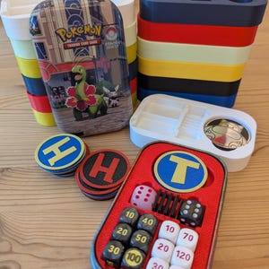May include: A Pokemon Trading Card Game metal tin featuring Meganium and Snivy, stacked plastic storage trays in white, yellow, red, black, and blue, circular tokens with H and T letters, and a red tray insert containing dice and number cubes.