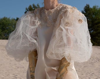 May include: A cream-colored wedding dress with sheer, voluminous sleeves and a ruffled skirt. The dress features gold-colored gloves and a detailed lace neckline. The background is a sandy beach under a clear blue sky.