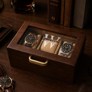 May include: A rectangular dark wood watch box with a glass lid featuring the white printed text "WALTER" and a large "W" monogram. The box has a gold-toned metal handle and contains two wristwatches on beige cushions inside the display case.