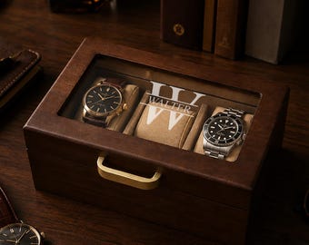 May include: A rectangular dark wood watch box with a glass lid featuring the white printed text "WALTER" and a large "W" monogram. The box has a gold-toned metal handle and contains two wristwatches on beige cushions inside the display case.