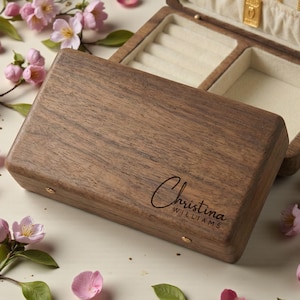 Puede incluir: Un joyero de madera con una inscripción personalizada, "Christina Williams", está abierto para revelar un interior color crema. La caja está rodeada de flores y pétalos rosas. El joyero es de madera oscura.