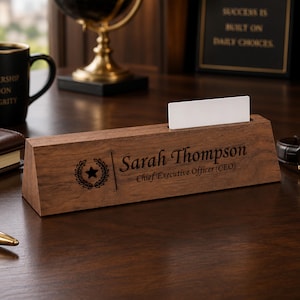 Puede incluir: Una placa de escritorio triangular de madera con el nombre grabado Sarah Thompson y el título Chief Executive Officer (CEO) junto a un emblema circular de corona de laurel con una estrella en el centro, incluyendo una ranura con una tarjeta blanca.