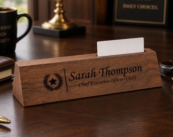 May include: A triangular wooden desk nameplate featuring the engraved name Sarah Thompson and the title Chief Executive Officer (CEO) alongside a circular laurel wreath emblem with a star in the center, including a slot holding a blank white card.