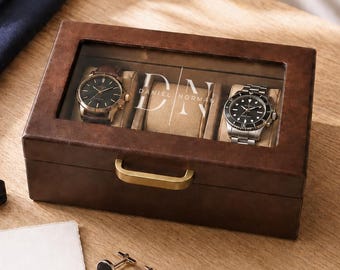 May include: A rectangular brown watch box with a glass lid featuring the white text DANIEL NORMAN. Inside are two wristwatches on beige cushions. Beside the box are a black bow tie, a white envelope with a gold wax seal, and two silver cufflinks.