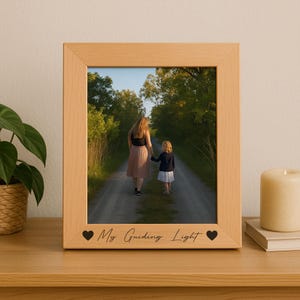 May include: A light-colored wooden picture frame with a photograph of a person and a child walking down a path. The frame has the text "My Guiding Light" with heart symbols. The frame is sitting on a wooden surface.