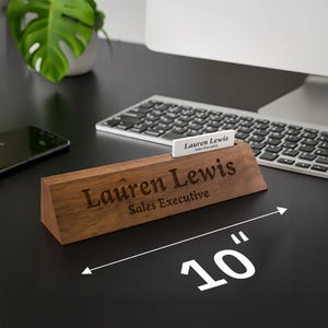 Puede incluir: Un letrero de escritorio de madera, de 25 cm de largo, con el nombre "Lauren Lewis" y el t&iacute;tulo "Sales Executive" grabados. Una tarjeta de visita con el mismo nombre y t&iacute;tulo est&aacute; en una ranura. El letrero est&aacute; en un escritorio negro.