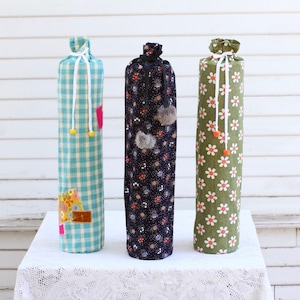 May include: Three fabric-covered storage containers with drawstring closures. The containers are blue and white gingham, black with white floral print, and green with white daisy print.