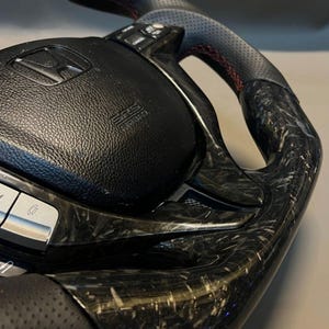 May include: A close-up of a black and carbon fibre steering wheel. The wheel has a perforated leather grip with red stitching. The centre features the Honda logo and SRS airbag label.