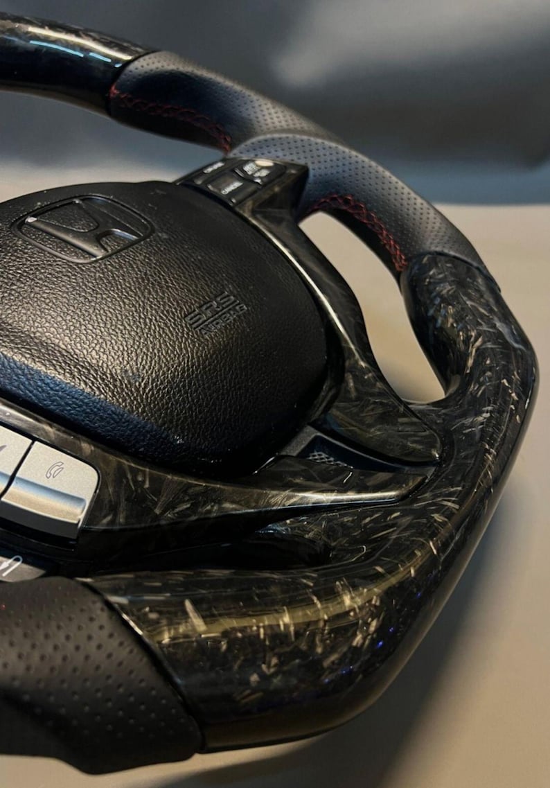 May include: A close-up of a black and carbon fibre steering wheel. The wheel has a perforated leather grip with red stitching. The centre features the Honda logo and SRS airbag label.