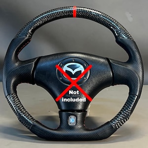 Puede incluir: Volante negro y de fibra de carbono con una franja roja en la parte superior. El volante tiene un logotipo de Mazda en el centro y el texto "Not included" encima. Tiene un agarre de cuero perforado y costuras rojas.