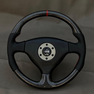 May include: A black and carbon fibre steering wheel with a red stripe at the top. The wheel has a black centre with the "MOMO" logo and red stitching accents. The wheel is designed for a vehicle.