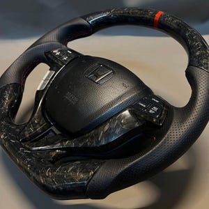 May include: A black and carbon fibre steering wheel with a red accent stripe. The wheel has perforated leather grips and integrated controls. The centre hub displays the Honda logo and SRS Airbag text.
