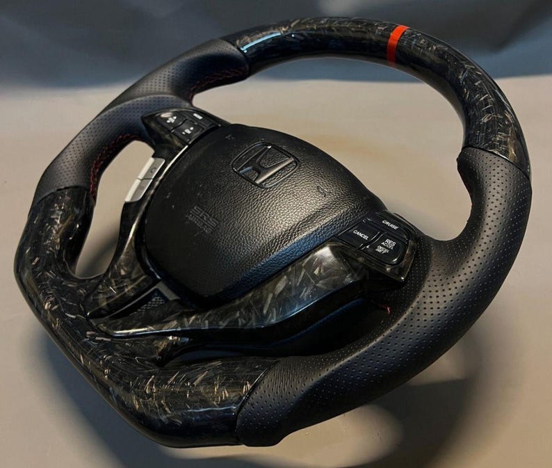 May include: A black and carbon fibre steering wheel with a red accent stripe. The wheel has perforated leather grips and integrated controls. The centre hub displays the Honda logo and SRS Airbag text.