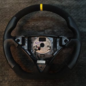 Puede incluir: Volante negro con empuñadura de cuero perforado y una franja amarilla en la parte superior. El volante tiene una textura similar a la gamuza en las zonas de agarre. El cubo central es negro con varios puntos de montaje.
