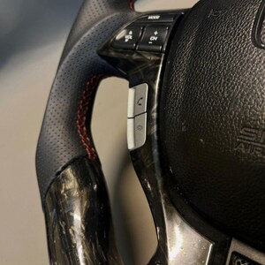 May include: Close-up of a black and carbon fibre steering wheel with red stitching. The wheel has integrated controls for mode, volume, and phone functions. The design includes a perforated leather grip for enhanced handling.