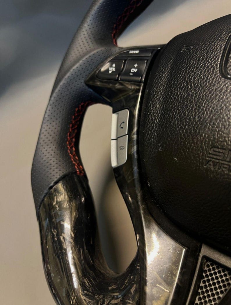 May include: Close-up of a black and carbon fibre steering wheel with red stitching. The wheel has integrated controls for mode, volume, and phone functions. The design includes a perforated leather grip for enhanced handling.