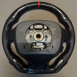 May include: A black and carbon fibre steering wheel with red stitching and a red stripe at the top. The wheel has a perforated leather grip and a central hub with exposed components.