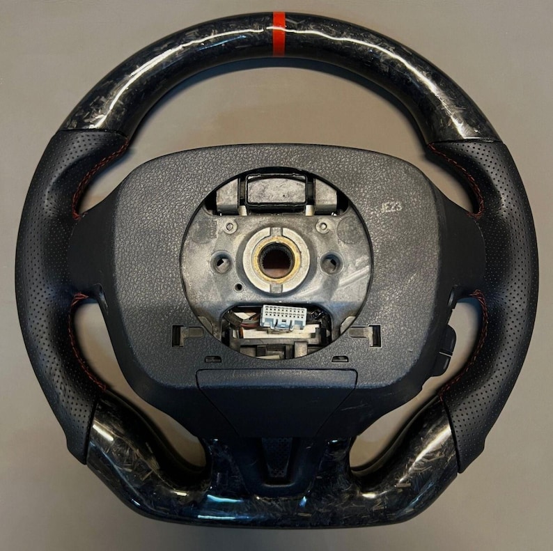 May include: A black and carbon fibre steering wheel with red stitching and a red stripe at the top. The wheel has a perforated leather grip and a central hub with exposed components.