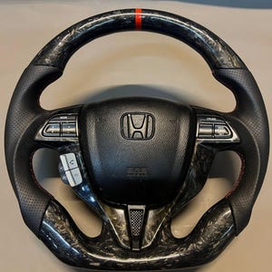 May include: Black and carbon fibre steering wheel with red stitching and a red stripe at the top. The centre features the Honda logo and control buttons. The wheel has a perforated leather grip.