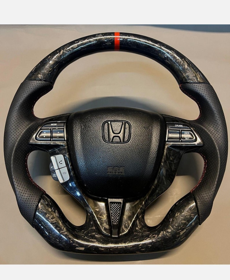 May include: Black and carbon fibre steering wheel with red stitching and a red stripe at the top. The centre features the Honda logo and control buttons. The wheel has a perforated leather grip.