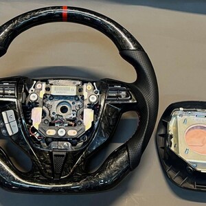 May include: A black and carbon fibre steering wheel with red accent stitching and a red stripe at the top. The image also shows a detached airbag module.