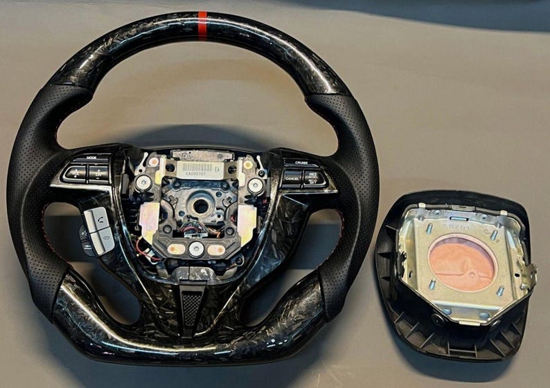 May include: A black and carbon fibre steering wheel with red accent stitching and a red stripe at the top. The image also shows a detached airbag module.