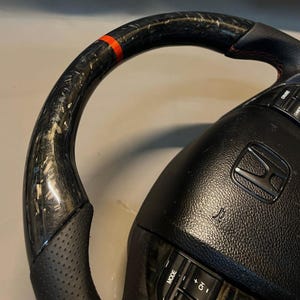 May include: Close-up of a black and carbon fibre steering wheel with a red accent stripe. The wheel has a perforated leather grip and integrated controls. The Honda logo is visible in the centre.
