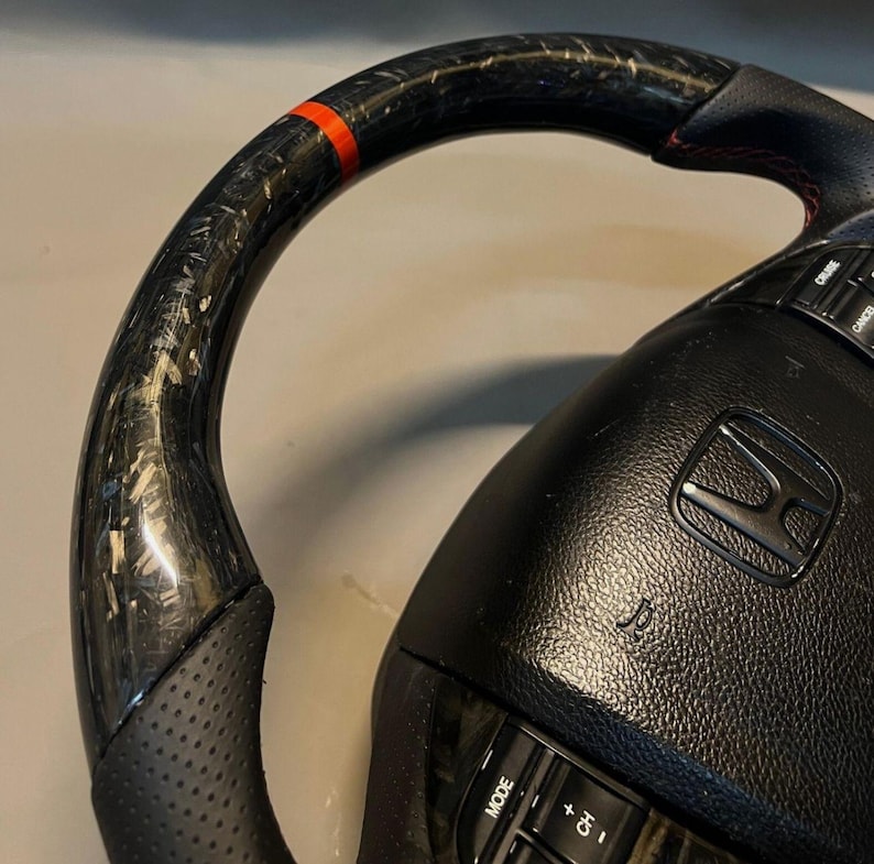 May include: Close-up of a black and carbon fibre steering wheel with a red accent stripe. The wheel has a perforated leather grip and integrated controls. The Honda logo is visible in the centre.