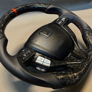 May include: Black and carbon fibre steering wheel with red stitching and a red stripe. The wheel features integrated controls and the Honda logo. The wheel has a perforated leather grip.