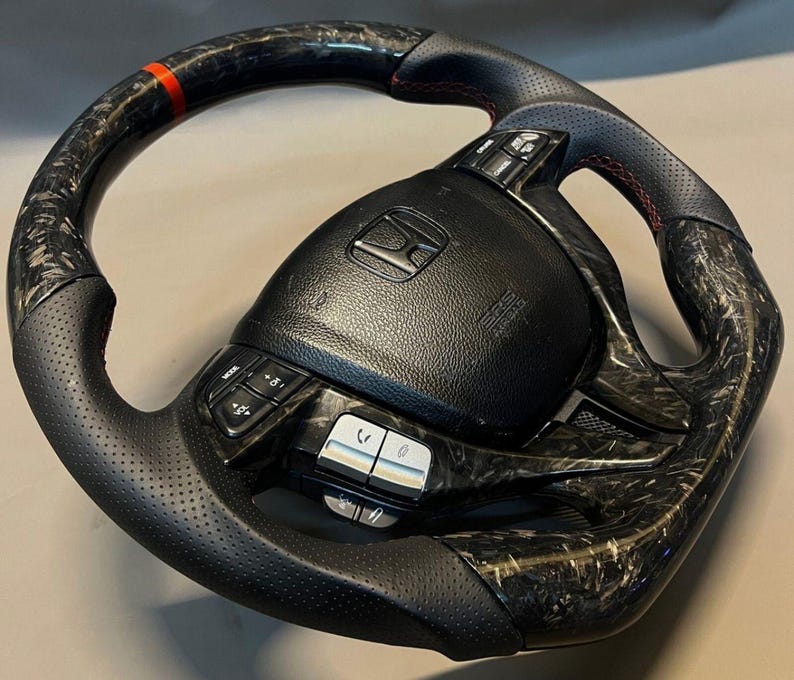 May include: Black and carbon fibre steering wheel with red stitching and a red stripe. The wheel features integrated controls and the Honda logo. The wheel has a perforated leather grip.