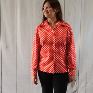 May include: A red button-down shirt with white polka dots. The long-sleeved shirt is paired with black pants and tan sandals. The outfit is worn by a person standing in front of a white wooden background.