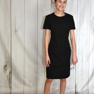 May include: A young person wearing a black short-sleeved dress with a textured pattern. The dress has a round neckline and a straight silhouette. The person is standing in front of a white wood panel background.
