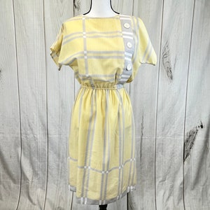 Peut inclure: Une robe à carreaux jaunes et gris avec des manches courtes et un détail boutonné sur le côté. La robe a un corsage ajusté et une jupe ample.