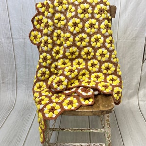 May include: A crocheted throw blanket with a yellow and brown floral pattern. The blanket is folded and draped over a wooden chair.