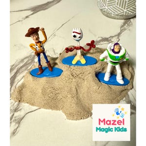 May include: Three Toy Story character figurines, Woody, Forky, and Buzz Lightyear, stand on blue platforms in a sand-like substance. The Woody figure wears a cowboy hat and vest. The Forky figure has red arms. The Buzz Lightyear figure is in a green and white spacesuit.
