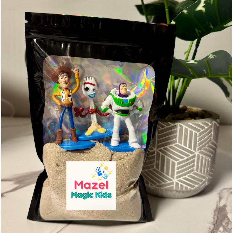 May include: A black bag with a clear window containing three Toy Story character figurines: Woody, Forky, and Buzz Lightyear. The bag also contains sand and a label that reads "Mazel Magic Kids."
