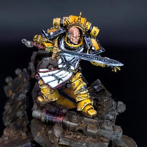 Puede incluir: Figura en miniatura de un guerrero con armadura amarilla y negra, sosteniendo una espada plateada. La figura está sobre una base rocosa detallada con rayas de peligro amarillas y negras. La espada tiene el texto "IMPERATORI".