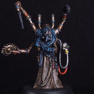 May include: A miniature figurine of a robed figure with mechanical arms and a skull-like mask. The figure is painted in shades of brown, blue, and gold, with intricate details. It holds a saw blade and a knife, with tubes and wires attached.