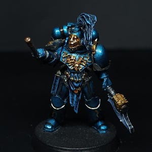 Puede incluir: Figura en miniatura de un marine espacial con armadura azul y detalles dorados. La figura está de pie sobre una base negra y sostiene un arma. La armadura tiene detalles intrincados y un brillo metálico.