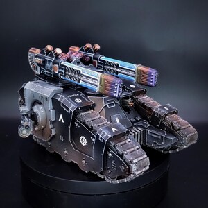 May include: A detailed miniature tank model, predominantly black with metallic accents. The model features two large, multi-barreled cannons with blue and purple highlights. The tank's treads and body have intricate detailing, and the word "VEHICLE" is visible.