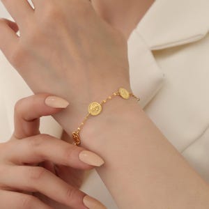 Peut inclure: Un bracelet doré orné de plusieurs breloques circulaires. Les breloques présentent un motif détaillé et le bracelet est délicat. Le bracelet est porté au poignet, et le teint est clair. L'arrière-plan est de couleur claire.