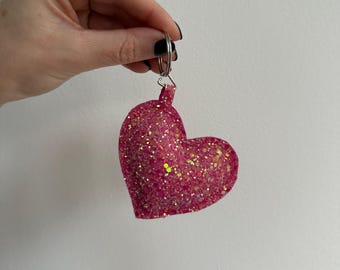 May include: A pink heart-shaped keychain with glitter. The keychain is held by a hand against a white background. The heart is a vibrant pink color with gold glitter accents, and a silver keyring.