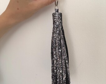 May include: A silver glitter tassel keyring with a black backing and a silver metal ring. The tassel is made of multiple strips of glittery material, creating a shimmering effect. The keyring is held against a white background.