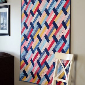 May include: A colorful quilt with a woven pattern in shades of blue, pink, red, and yellow. The quilt is hanging on a white wall next to a white chair.