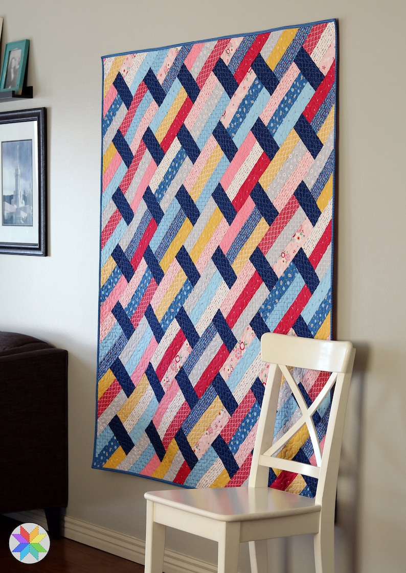 May include: A colorful quilt with a woven pattern in shades of blue, pink, red, and yellow. The quilt is hanging on a white wall next to a white chair.