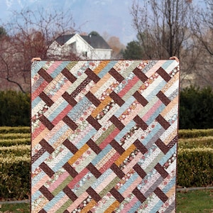 May include: A brown, pink, blue, and yellow patchwork quilt with a chevron pattern. The quilt is made of various floral fabrics and is displayed in front of a mountain and a house.