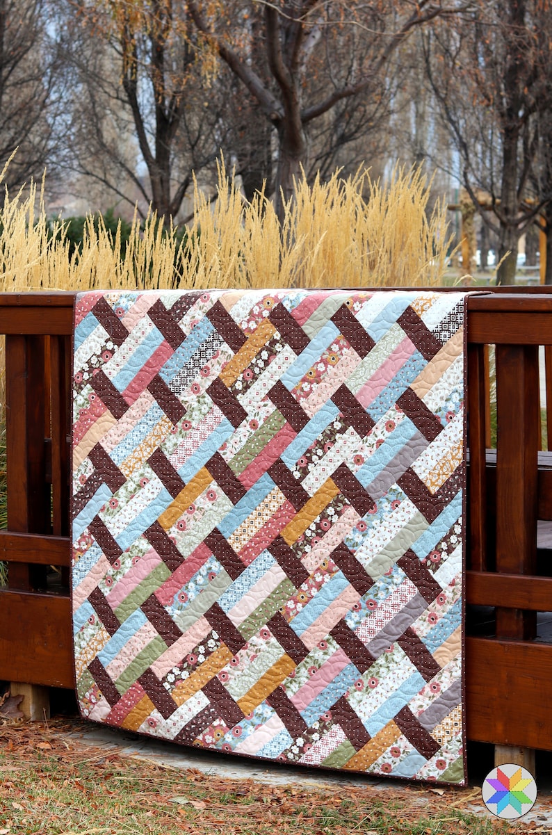 May include: A brown, pink, blue, and yellow patchwork quilt with a chevron pattern. The quilt is hanging on a wooden fence.