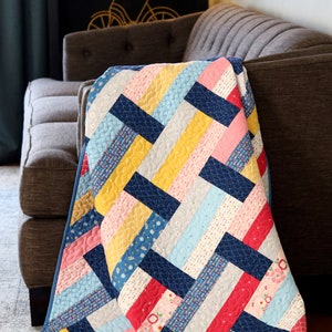 May include: A colorful quilt with a chevron pattern in shades of blue, yellow, pink, and red. The quilt is made of various patterned fabrics and is folded on a gray couch.