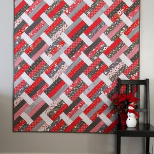 May include: A red, white, and gray patchwork quilt with a geometric pattern. The quilt is hanging on a gray wall and is framed by a white baseboard.